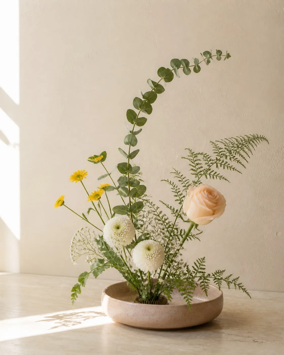 A sunny Ikebana-style arrangement of a peach rose, white chrysanthemums, yellow daisies, baby's breath, eucalyptus and fern foliage in a shallow ceramic bowl with a visible kenzan